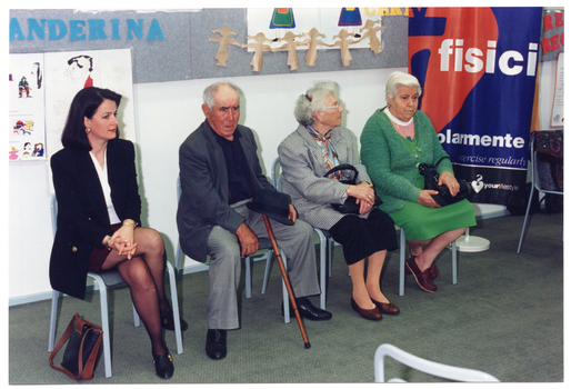 COL photo of four people who are seated on a row of chairs. The man has a walking stick leaning against his leg. On the wall behind them are several posters, and beside them is a colourful frees-standing banner.
