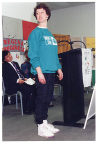 COL photo of a woman standing at a black lectern. She is standing straight and has her head turned to one side. Behind her are a few people, (seated and partially obscured), who also have their heads turned to one side. There are several posters on the wall behind them.