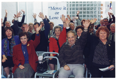 COL photo of a large group of people who are seated on rows of chairs. They are each holding one arm up, outstretched, with their other hand on their laps. Behind them, there is a freestanding "Move It Or Lose It" banner.