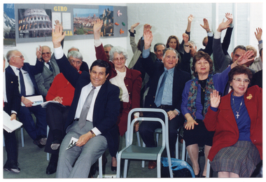 COL photo of a large group of people who are seated on rows of chairs. They are each holding one arm up, outstretched, with their other hand on their laps. On the wall are several Italian travel posters.