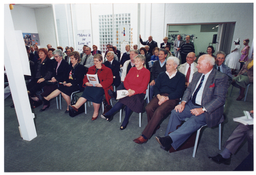 COL photo of a group of people, seated, stretching one leg forward while lifting it off the ground. Behind them are some people standing in the background. Two of these people appear to be in costume. On the wall are some travel posters, and at the back of the room is a freestanding "Move It Or Lose It" banner.