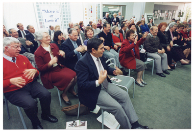 COL photo of a large group of people, seated. They are shaking their hands as a warm-up exercise. There are some printed brochures on some chairs and on the floor. In the background is a freestanding "Move It Or Lose It" banner beside an open doorway, through which some people are standing. Beside them are two people who appear to be in costume.