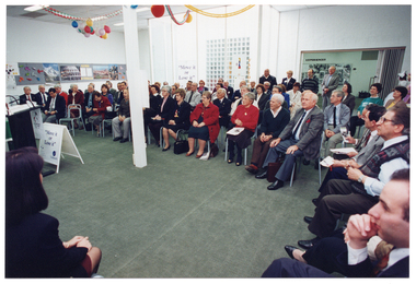 COL photo of a large group of people who are seated in a hall, with a lectern in front of them. In the foreground, there is a woman (partially obscured). In front of the lectern, there is a "Move It Or Lose It" A-frame sandwich board and a "Move It Or Lose It" freestanding banner in the background. There are travel posters on the wall. Some people are standing at the back, two of whom appear to be in costume.