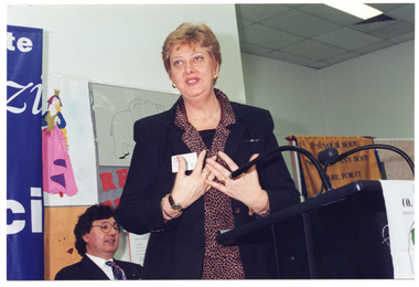 COL medium close-up photo of a woman standing at a black lectern, mid-speech. Behind her, a man is seated (partially obscured). There are several posters on the wall behind them.