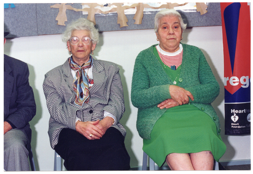 COL photo of three people (one partially obscured) seated on chairs against the wall. Behind them, there is a display board on the wall.
