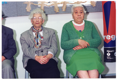 COL photo of three people (one partially obscured) seated on chairs against the wall. Behind them, there is a display board on the wall.