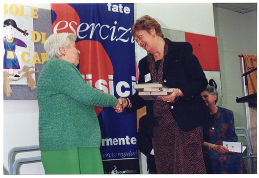 COL photo of a woman holding a small stack of wrapped gifts as she shakes the hand of an elderly woman. Behind them is a woman (seated) and a colourful freestanding banner.