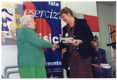 COL photo of a woman holding a small stack of wrapped gifts as she shakes the hand of an elderly woman. Behind them is a woman (seated) and a colourful freestanding banner.