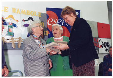 COL photo of a woman holding a small stack of wrapped gifts as she shakes the hand of an elderly woman, while another woman looks on. Behind them is a woman (seated), a colourful freestanding banner, and a display board.