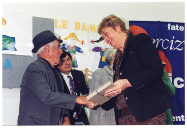 COL photo of a woman holding a wrapped gift as she shakes the hand of an elderly man, while another man looks on. Beside them is a woman (partially obscured), a colourful freestanding banner, and a display board.