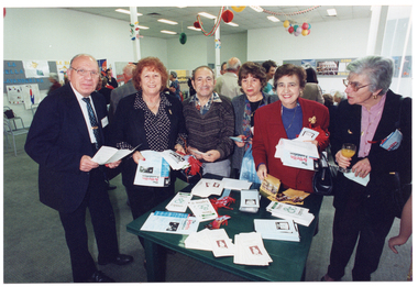 COL photo of a small group of people gathered around a display table. There is a range of printed brochures on top of the table, which they are perusing. In the background, there is a small group of people, also standing. There are posters on display boards around the room. Balloons, streamers, and paper decorations are hanging from the ceiling.