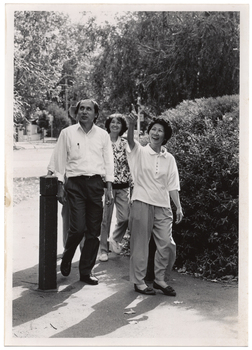 B&W photo of a small group of people taking a walk along a concrete path. There is a wooden bollard across the entrance to the path. Beside the path is a shrub, and there are some tall trees in the background.
