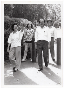 B&W photo of a small group of people taking a walk along a concrete path. One woman is looking up and pointing. There are two wooden bollards across the entrance to the path. Beside the path are some large shrubs, and there are some tall trees in the background.