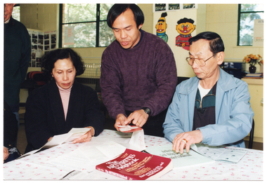 COL photo of a small group of people, seated and standing, gathered around a table. They are looking at some brochures. Also on the table is a book titled "The Arthritis Helpbook" and a plastic display folder with a clear front cover. Printed on the cover page inside the folder is the text, "Arthritis Self Help Course Leaders Manual". In the background, there are some large windows with a view to some trees outside. There are some storage cupboards along the wall beneath the windows. There are various pictures on the walls, including photos and a large cartoon drawing of Bert and Ernie (two characters from the USA children's television show, Sesame Street).