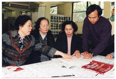 COL photo of a small group of people around a rectangular table. One person is standing while the others are all seated. They are looking at some brochures. One woman is pointing to some content in a brochure. Also on the table are a plastic display folder and a book titled, "The Arthritis Helpbook".