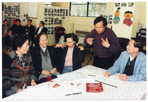 COL photo of a small group of people around a rectangular table. One person is standing while the others are all seated. Two people are looking down at a brochure and practicing the hand exercises illustrated. On the table, there are several brochures, a plastic display folder, and a book titled, "The Arthritis Helpbook". In the background, there are a few people sitting on some chairs against the wall.