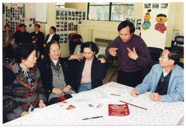 COL photo of a small group of people around a rectangular table. One person is standing while the others are all seated. Two people are looking down at a brochure and practicing the hand exercises illustrated. On the table, there are several brochures, a plastic display folder, and a book titled, "The Arthritis Helpbook". In the background, there are a few people sitting on some chairs against the wall.