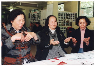 COL photo of three women sitting at a table. They are practising some hand exercises. There are several brochures on the table. In the background, there are a few people (partially obscured) sitting on chairs near the wall and around the room. The arm of one of the people in the background is in an orthopaedic splint.