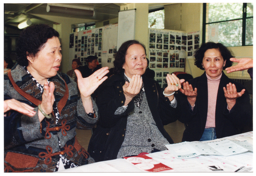 COL photo of three women sitting at a table. They are practising some hand exercises. There are several brochures on the table. In the background, there are a few people (partially obscured) sitting on chairs near the wall and around the room.