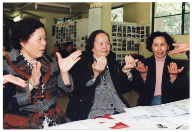 COL photo of three women sitting at a table. They are practising some hand exercises. There are several brochures on the table. In the background, there are a few people (partially obscured) sitting on chairs near the wall and around the room.