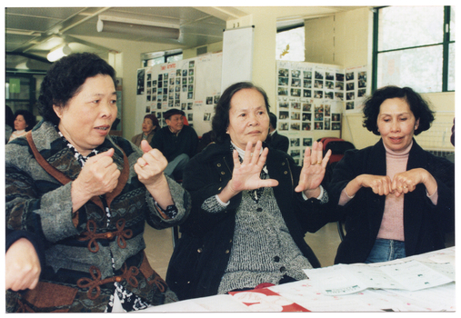 COL photo of three women sitting at a table. They are practising some hand exercises. There are several brochures on the table. In the background, there are a few people (partially obscured) sitting on chairs near the wall and around the room.