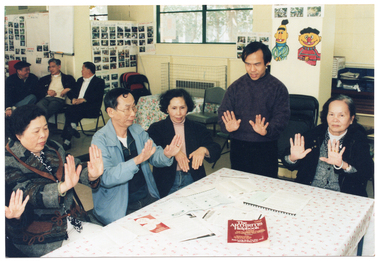 COL photo of a small group of people gathered around a rectangular table. One person, who is standing, demonstrates some hand exercises. The others, who are seated, are also doing the hand exercises. On the table, there are brochures, a plastic display folder, and a book titled, "The Arthritis Helpbook". In the background, there are a few people sitting on some chairs against the wall.