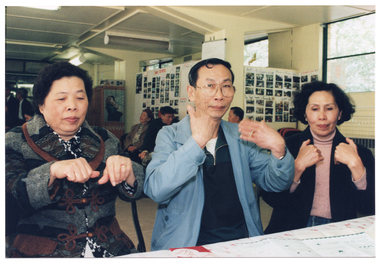 COL photo of two women and a man sitting at a table. They are practising some hand exercises. There are several brochures on the table. In the background, there are a few people (some partially obscured) either sitting on chairs or standing.