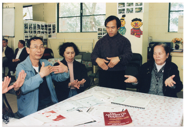 COL photo of a small group of people gathered around a rectangular table. One person, who is standing, demonstrates some hand exercises. The others, who are seated, are also doing the hand exercises. On the table, there are brochures, a plastic display folder, and a book titled, "The Arthritis Helpbook". In the background, there are a few people sitting on some chairs against the wall.