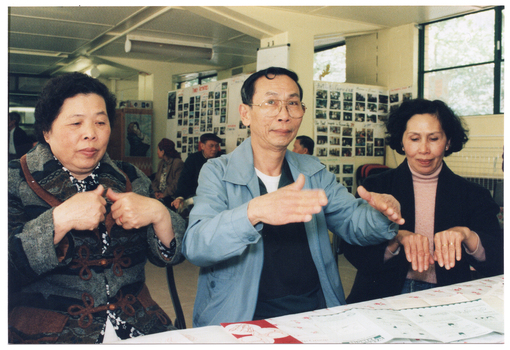 COL photo of two women and a man sitting at a table. They are practising some hand exercises. There are several brochures on the table. In the background, there are a few people (some partially obscured) either sitting on chairs or standing.