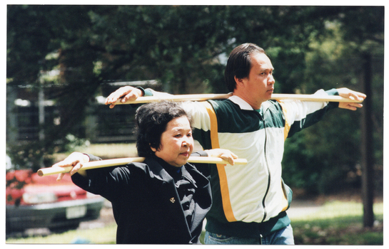 COL photo of two people exercising outdoors. They are using wooden poles as part of their exercise routine. In the background, there are several large trees and a red car (partially obscured).