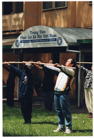 COL photo of a group of people exercising with wooden poles, on a grass lawn outdoors. They are holding the poles horizontally at shoulder height, with their arms outstrectched in front of them. Behind them is a brick building, with a sign that reads "Trung Tâm Phúc Loi Xã Hôi Viêt Nam Vietnamese Welfare Resource Centre A program of Jesuit Social Services", with a logo on either side of the text.