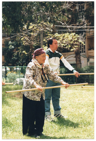 COL photo of a man and an elderly woman exercising outdoors on a grass lawn. They each are holding a wooden pole horizontally in front of them, at waist height. In the background, there is a multi-story building in the distance.