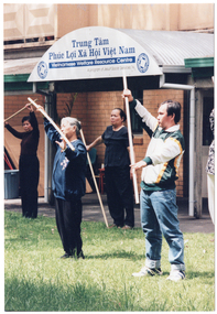 COL photo of a group of people exercising with poles, on a grass lawn outdoors. They are holding the poles diagonally, with their arms outstrectched in front of them. Behind them is a brick building, with a sign that reads "Trung Tâm Phúc Loi Xã Hôi Viêt Nam Vietnamese Welfare Resource Centre A program of Jesuit Social Services", with a logo on either side of the text.