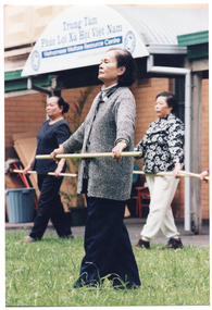 COL photo of three women exercising on a grass lawn outdoors with poles, which they are holding in front of them at waist height. Behind them is a brick building, with a sign which reads "Trung Tâm Phúc Loi Xã Hôi Viêt Nam Vietnamese Welfare Resource Centre.  The following text on the sign, "A program of Jesuit Social Services", and a logo on either side of the text, are blurred in this photo.