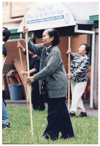 COL photo of a group of people (some partially obscured) exercising with poles on a grass lawn outdoors. They are holding the poles vertically in front of them. Behind them is a brick building, with a sign which is blurred. As per other photos in this series, the sign reads "Trung Tâm Phúc Loi Xã Hôi Viêt Nam Vietnamese Welfare Resource Centre A program of Jesuit Social Services", with a logo on either side of the text.