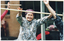 COL photo of an elderly woman (and two others, partially obscured) exercising outdoors with poles. They are holding the poles horizontally in front of their bodies, at head height. Behind them is a brick building (blurred).