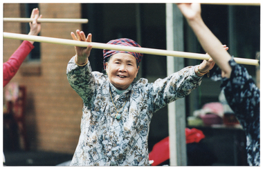 COL photo of an elderly woman (and two others, partially obscured) exercising outdoors with poles. They are holding the poles horizontally in front of their bodies, at head height. Behind them is a brick building (blurred).