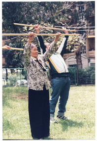 COL photo of a man and an elderly woman (and one other, partially obscured) exercising on a grass lawn outdoors with poles. They are holding the poles above their heads, with their arms outstretched. The man is leaning backwards, with his pelvis pushed forwards. There is a multi-story building in the distance behind them.