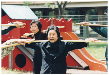 COL photo of a small group of people exercising outdoors with poles. They are stretching their arms across the poles, which are positioned horizontally behind their necks. Behind them is some playground equipment,  a fence, and a building.