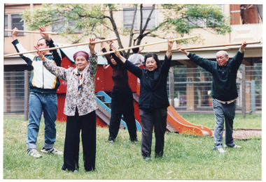 COL photo of a small group of people exercising with poles on a grass lawn outdoors. They are standing while holding the poles horizontally above their heads, with their arms outstreched. Behind them is some playground equipment, with a fence and a building in the background.