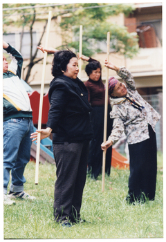 COL photo of a small group of people exercising with poles on a grass lawn outdoors. They are standing while holding the poles vertically behind them, with one arm pulled back behind their heads. Behind them is some playground equipment, with a fence and a building in the background.