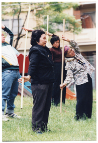 COL photo of a small group of people exercising with poles on a grass lawn outdoors. They are standing while holding the poles vertically behind them, with one arm pulled back behind their heads. Behind them is some playground equipment, with a fence and a building in the background.