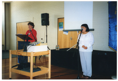 COL photo of two women standing at microphones on stands. One woman is standing behind a lectern. Hanging from the front of the lectern is an Arthritis Foundation of Victoria - Osteoporosis Victoria banner in blue and gold. In the space in front of and between them, there is a large wooden cart on castors. On it, there is a slide projector sitting on top of a white box. The projector is being propped up at an angle by a Yellow Pages telephone directory. It is facing a projector screen on a stand.