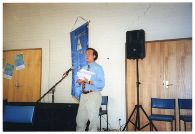 COL photo of a man speaking into a microphone on a stand. Behind him is a blue and gold "Arthritis Victoria - Osteoporosis Victoria" banner and a speaker on a stand. There are a few chairs pushed up against the wall and some closed double doors.