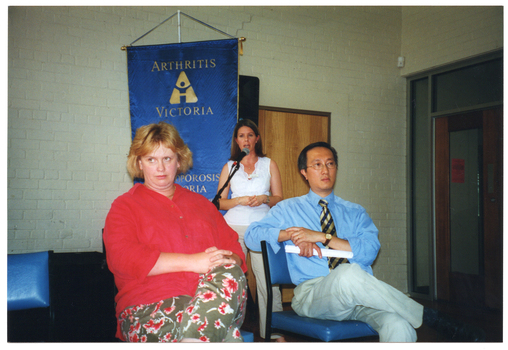 COL photo of a woman speaking into a microphone on a stand. Two other people are seated in front of her. Behind them is a blue and gold "Arthritis Victoria - Osteoporosis Victoria" banner.