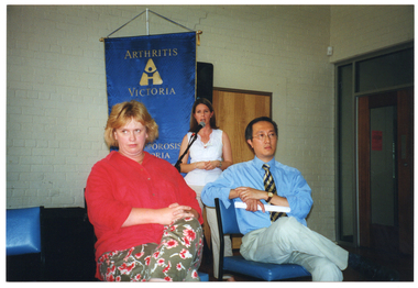 COL photo of a woman speaking into a microphone on a stand. Two other people are seated in front of her. Behind them is a blue and gold "Arthritis Victoria - Osteoporosis Victoria" banner.