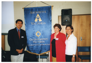 COL photo of three people standing beside a blue and gold Arthritis Victoria/Osteoporosis Victoria banner on a stand. Behind them, there is a speaker on a stand and a set of large double wooden doors.