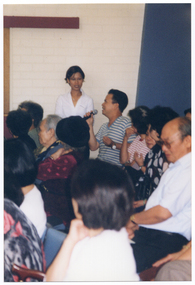 COL photo of a man speaking into a hand-held microphone. He is sitting amongst a large group of people on rows of chairs. A woman is standing next to him.