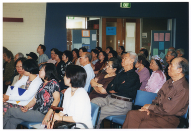 COL wide frame photo of a large group of people. They are seated on rows of chairs in a large room or hall. In the background, there are two freestanding vertical display boards on either side of a large set of double glass doors.