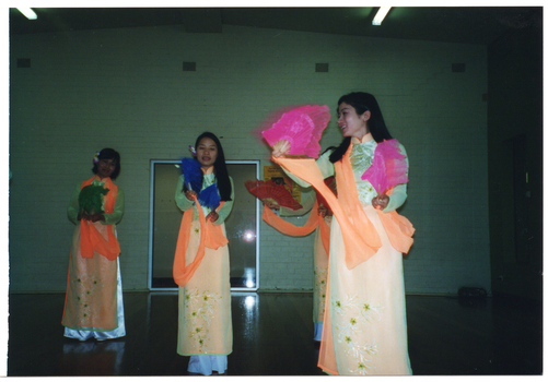 COL photo of four dancers (one partially obscured), wearing traditional Vietnamese dress. They are performing a dance with colourful hand-held fans.